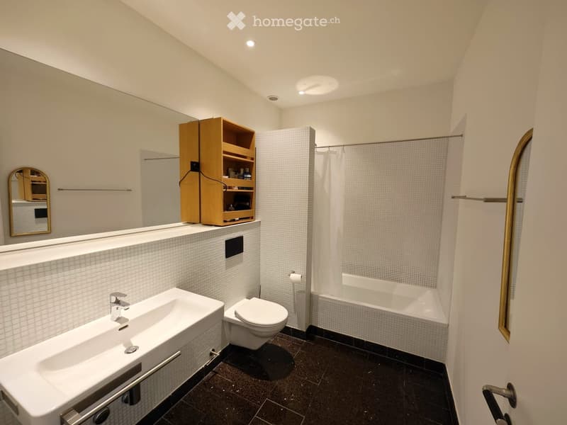 Modern bathroom with white tiled walls, dark tiled floor, sink, toilet, bathtub, and a wooden shelf above the sink.