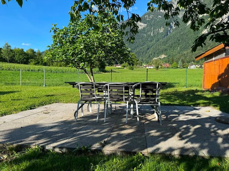Outdoor patio with a table and chairs, surrounded by a green lawn and a scenic mountain view.