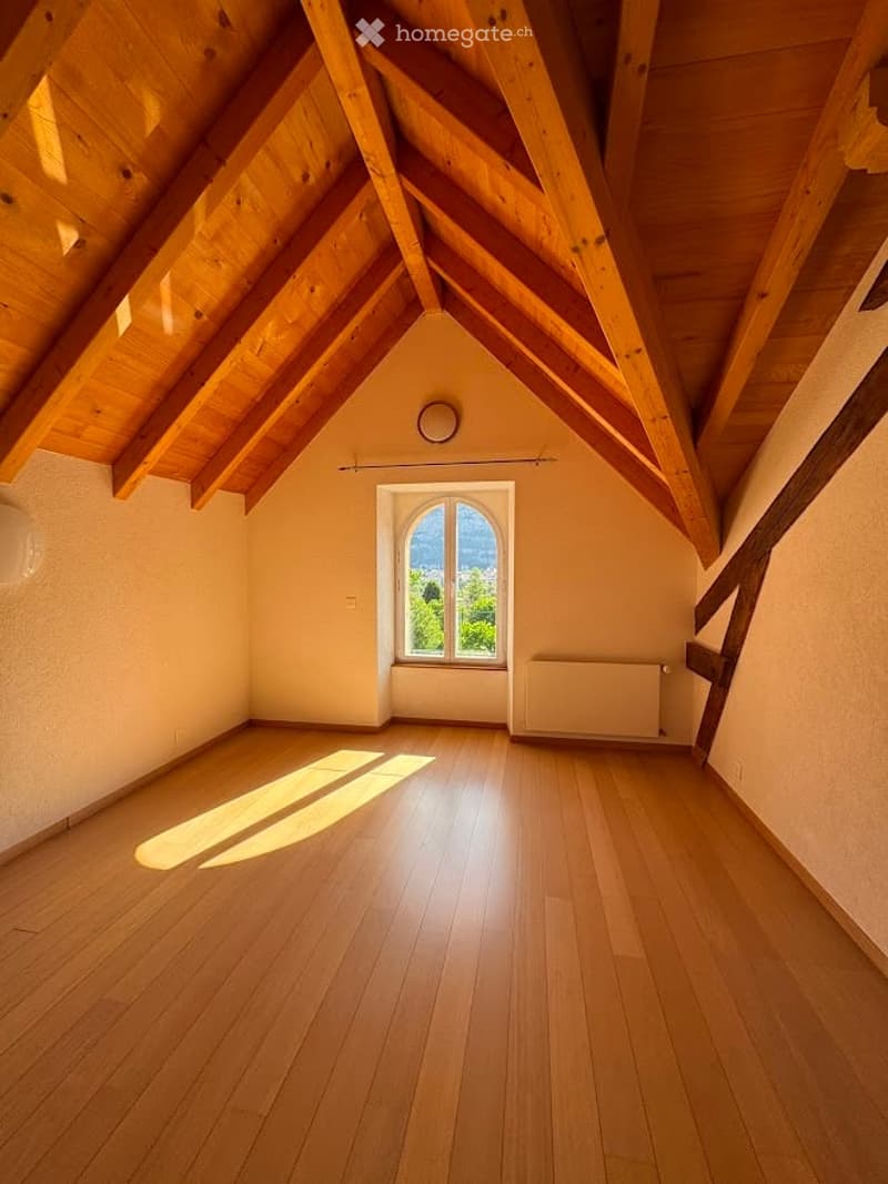 Bright, empty attic room with wooden floors, exposed wooden beams, and a large window offering a scenic view.