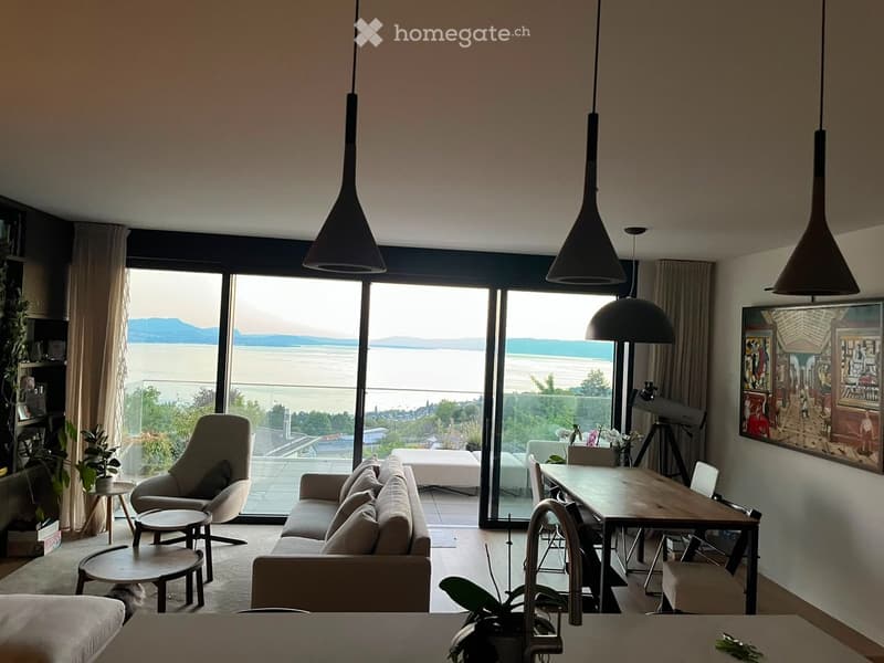 Modern living room with large windows offering a scenic view, featuring a sofa, chairs, a coffee table, pendant lights, and a painting on the wall.