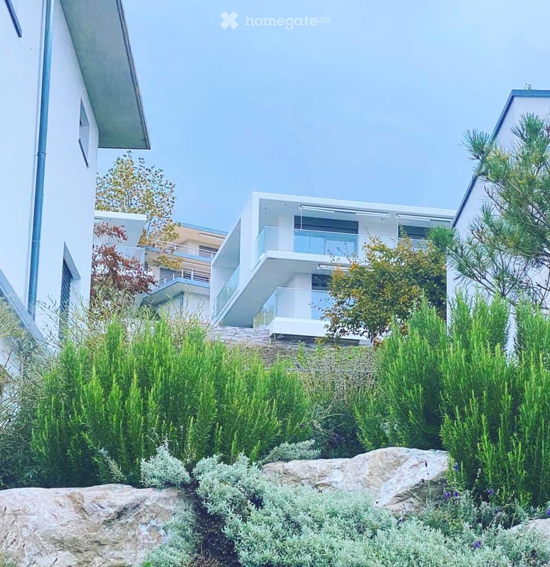 Modern residential complex with white buildings, glass balconies, and landscaped garden featuring shrubs, rocks, and trees.
