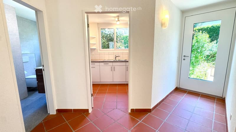 Hallway with terracotta tiles, leading to a kitchen and bathroom. Features a white door with glass panel and a wall-mounted light fixture.