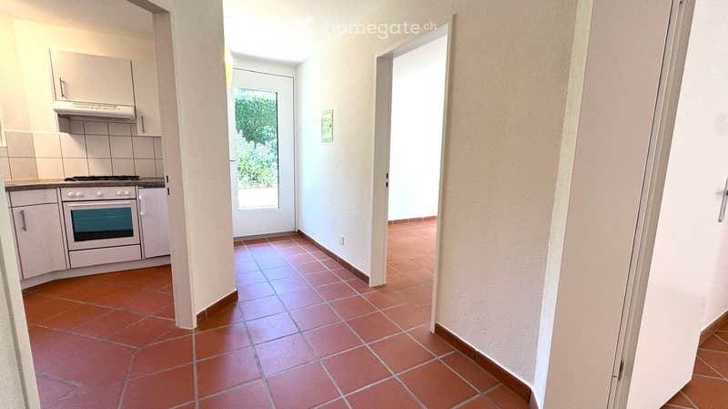 A narrow hallway with terracotta tiled flooring, white walls, and a view of a kitchen area with white cabinets, a stove, and an oven. The hallway leads to a door with a glass panel that opens to a green outdoor area.
