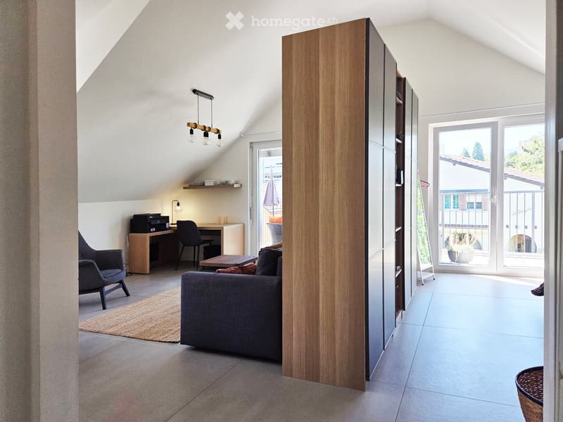 Modern living room with a grey sofa, armchair, coffee table, desk, and printer. Large windows and sliding doors lead to a balcony with outdoor furniture.