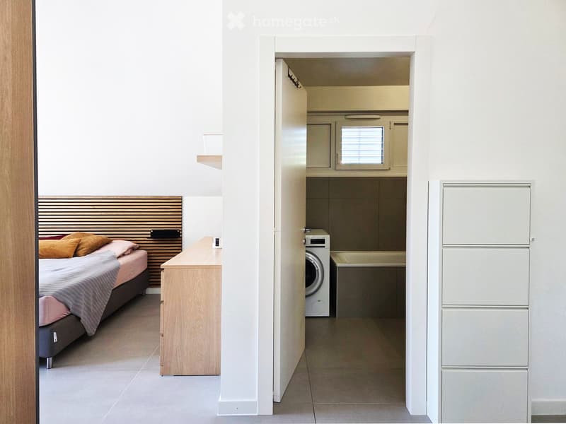 Modern, compact bedroom with a bed, nightstand, and an adjacent laundry area featuring a washing machine.