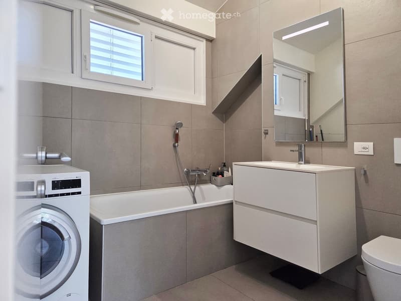 Modern bathroom with a bathtub, sink, mirror, toilet, and a private washing machine. Features include a window with blinds and sleek, contemporary fixtures.