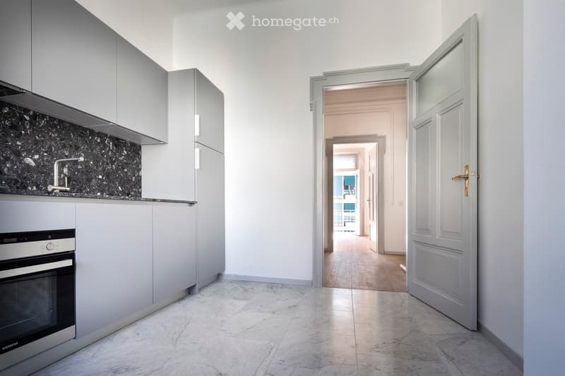Modern kitchen with grey cabinets, marble backsplash, and a Siemens oven. Open doorway leads to a hallway with wooden flooring.
