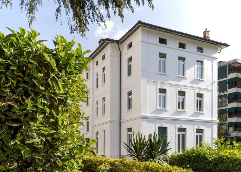 A white, multi-story building with classic architectural details, surrounded by greenery and trees.