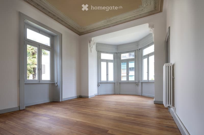 An empty room with wooden flooring, large windows, and decorative ceiling moldings. The space is well-lit with natural light and features a radiator on one wall.