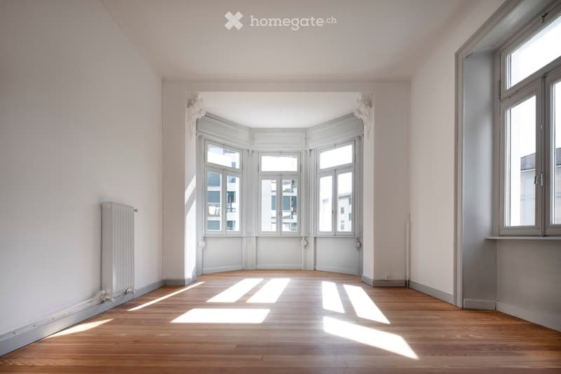 Empty room with wooden flooring, white walls, large windows, and a radiator on the left side.