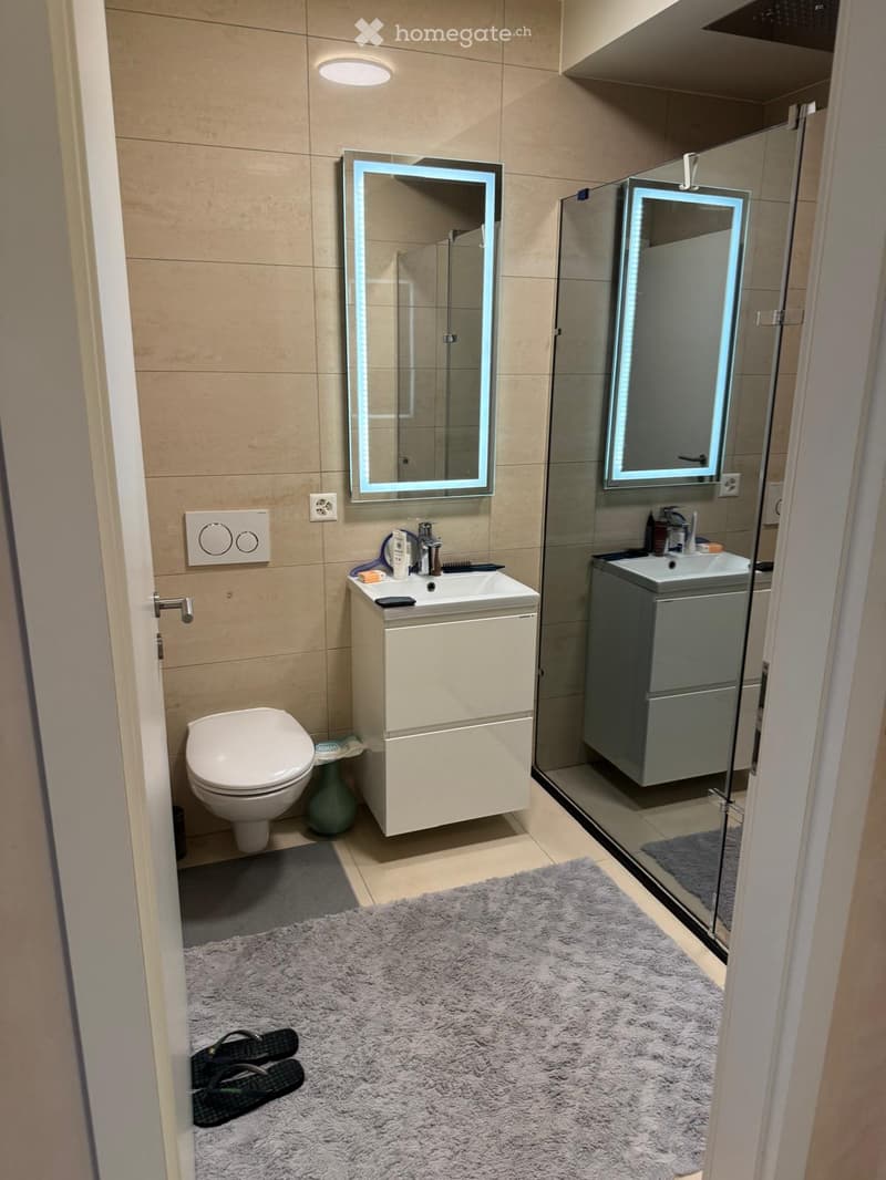 Modern bathroom with beige tiles, a toilet, a sink with a cabinet, a shower with a glass enclosure, and two illuminated mirrors above the sinks.