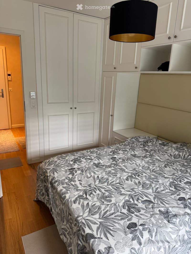 Bedroom with a bed covered in a grey and white leaf-patterned duvet, white built-in wardrobes, and a black pendant lamp with a gold base.