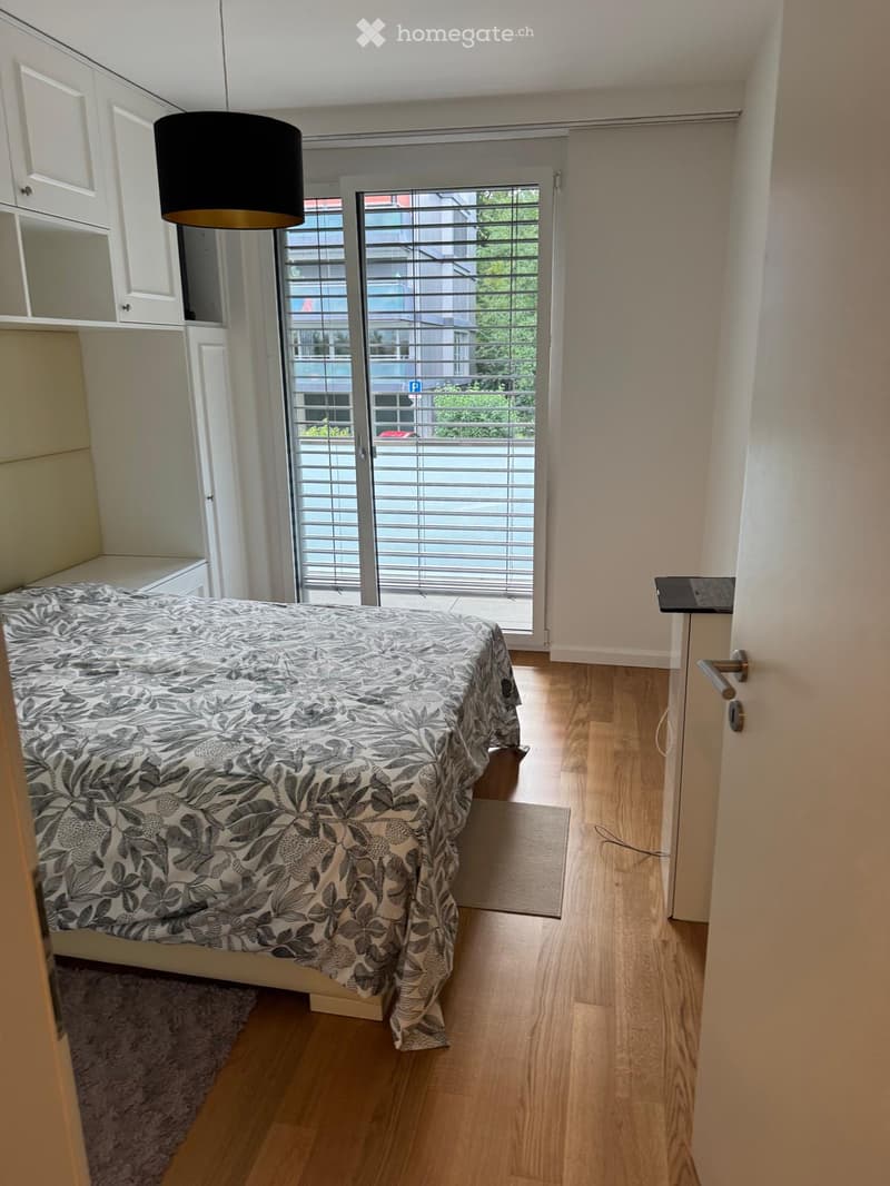 Modern bedroom with wooden flooring, a bed with a patterned bedspread, built-in white cabinets, and a pendant light fixture.