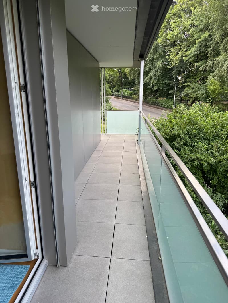 Modern balcony with tiled floor, glass railing, and view of greenery and street.