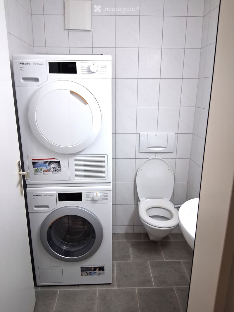 Small room with white tiled walls, white tiled floor, white toilet, white sink, stacked washing machine and dryer