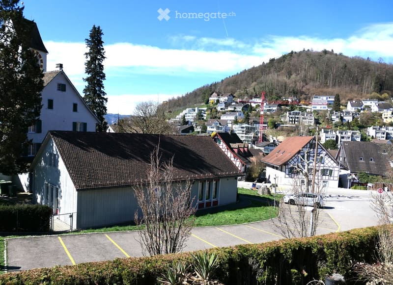 House with brown roof, white walls, located in a hilly area with a view of the town, surrounded by trees and bushes, with a parking lot in front.