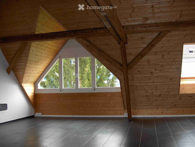 Empty attic space with wooden walls, exposed beams, and large windows offering a view of greenery outside.