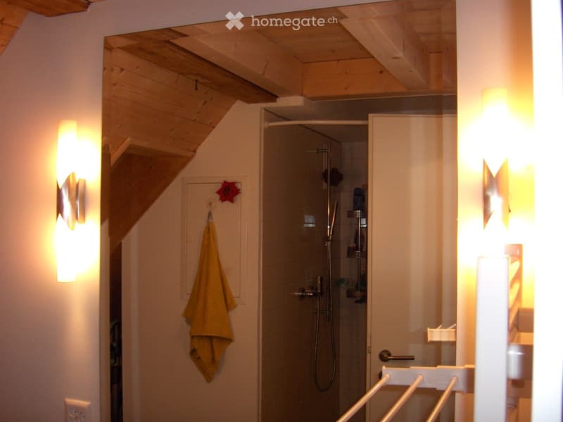 Modern bathroom with a glass-enclosed shower, a towel hanging on a rack, and built-in shelving for toiletries. The ceiling has exposed wooden beams, and the walls are painted white.