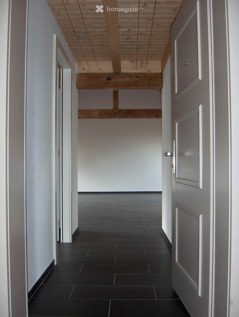 Empty hallway with dark tiled floor, white walls, and wooden ceiling beams. Doors visible on both sides.
