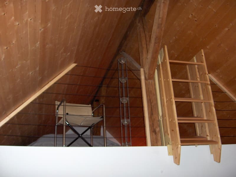 An attic space with wooden beams, a wooden ladder, and a metal chair, featuring exposed wooden ceiling and walls.