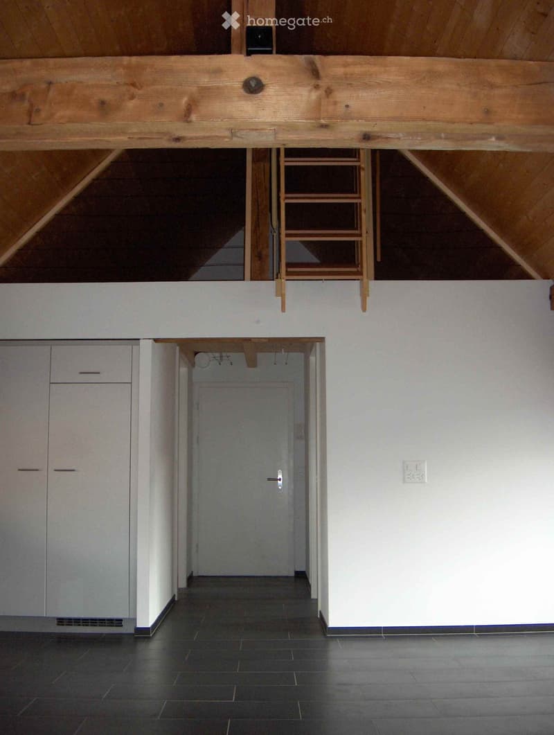 Empty hallway with dark tiled floor, white walls, and a door at the end. Exposed wooden beams and a small staircase lead to an upper level.