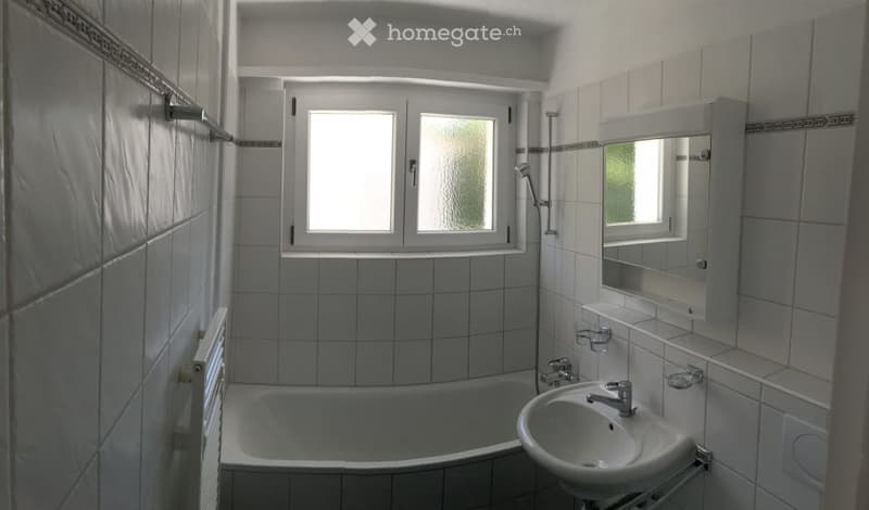 Bathroom with white tiled walls and floor, featuring a bathtub, sink, mirror, and window with frosted glass panels.