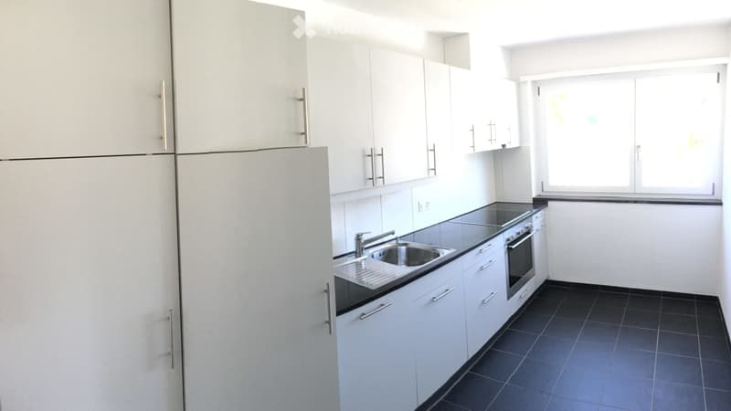 Modern kitchen with white cabinets, stainless steel sink, oven, and refrigerator. Dark tiled floor and large window.