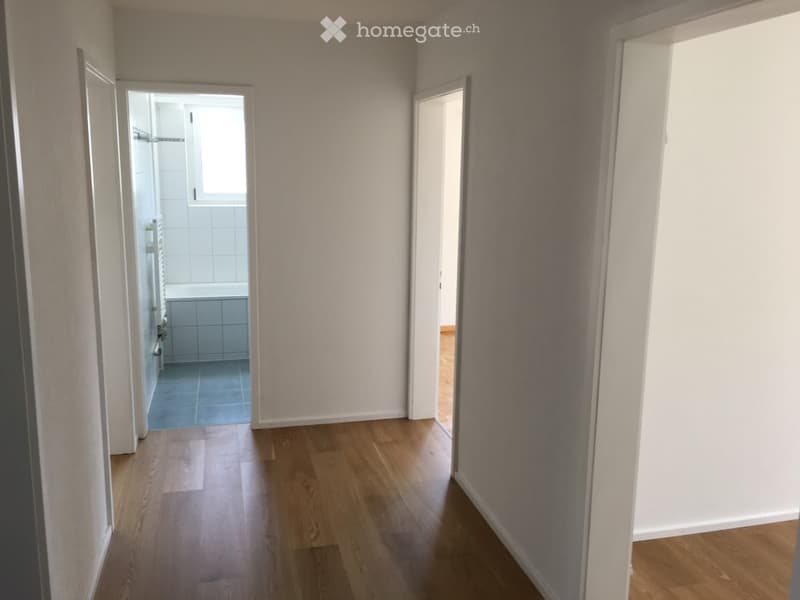 Empty hallway with wooden flooring and white walls, leading to a bathroom and another room.