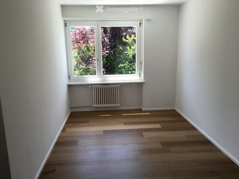 Empty room with wooden flooring, white walls, and a large window with a view of greenery outside. A radiator is visible below the window.