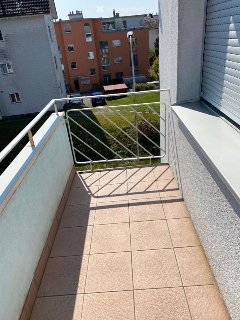 Balcony with tiled floor, metal railing, and view of a grassy area and neighboring buildings.