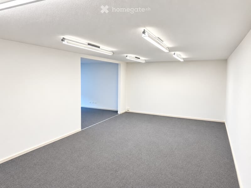Empty room with white walls, grey carpet, and recessed lighting. A doorway leads to another room with blue walls and carpet.