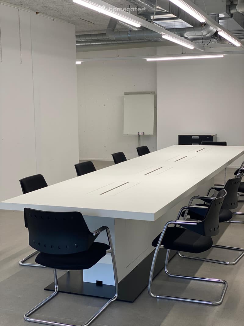 long desk, several black chairs, white wall, ceiling lamp