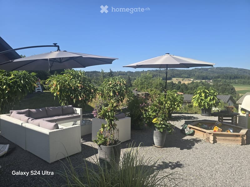 Outdoor patio with a white wicker sofa set, a wooden coffee table, two large umbrellas, and several potted plants. The area is surrounded by greenery and offers a scenic view of a valley.