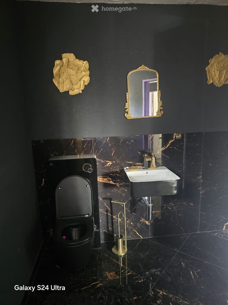 Modern bathroom with black marble walls and floor, featuring a sleek black toilet, a white sink with gold fixtures, and an ornate gold-framed mirror.