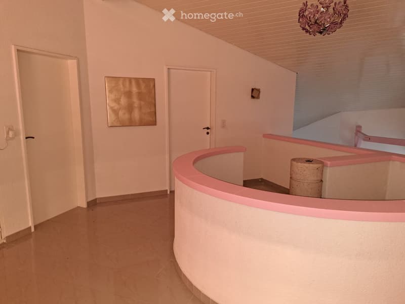 Empty hallway with light-colored walls, tiled floor, and a curved pink railing leading to a staircase. There are two doors and a decorative wall piece visible.