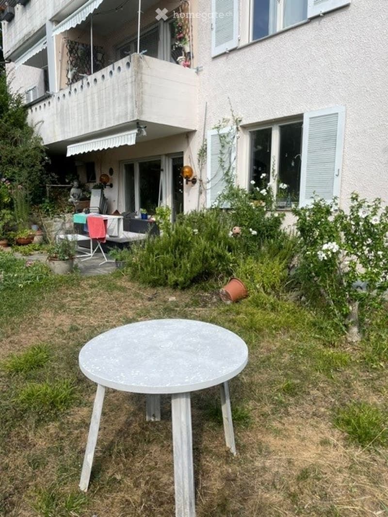 Garden area with a round table, potted plants, and a balcony with a covered seating area.
