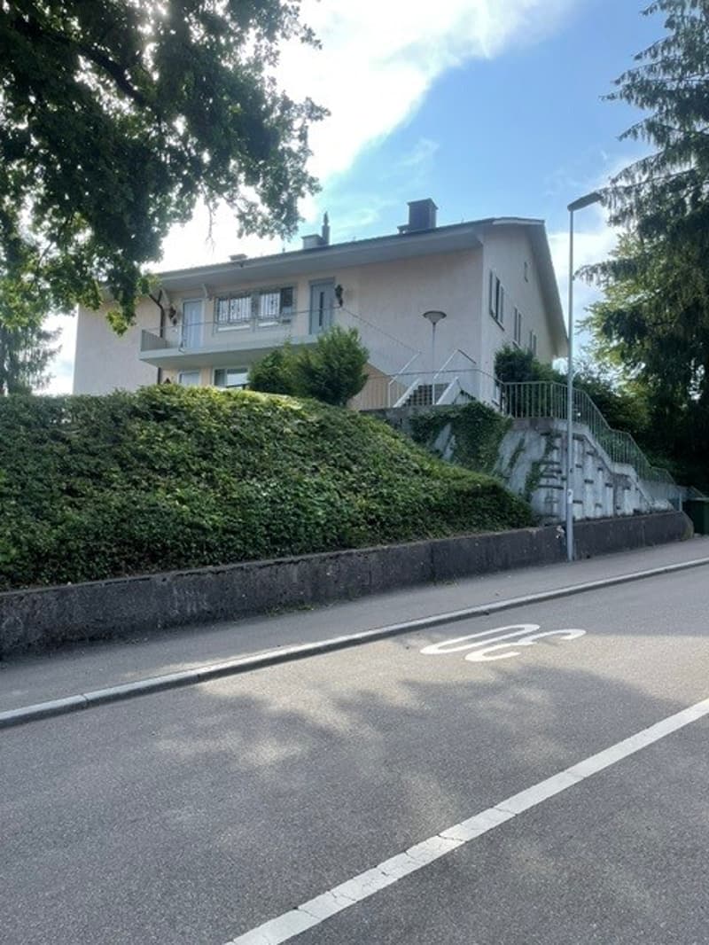 Two-story residential building with a raised ground floor, surrounded by greenery and a concrete retaining wall.