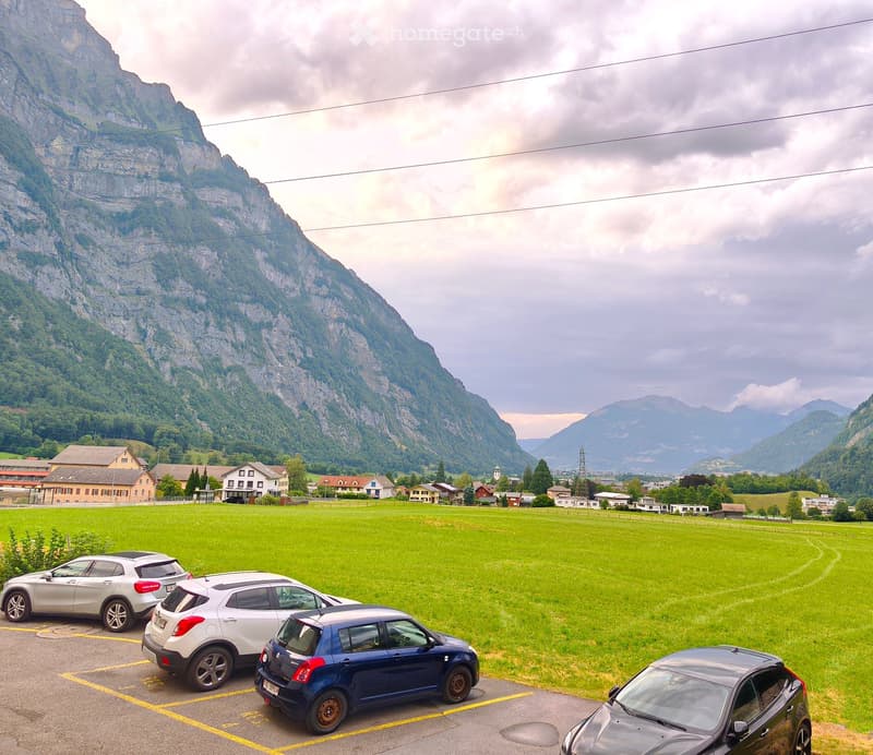 Parking lot with several cars, surrounded by a green field and distant mountains.