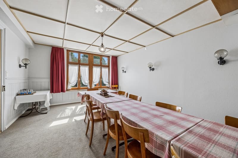 Dining room with wooden chairs and tables covered in plaid tablecloths, large windows with red curtains, and wall-mounted light fixtures.