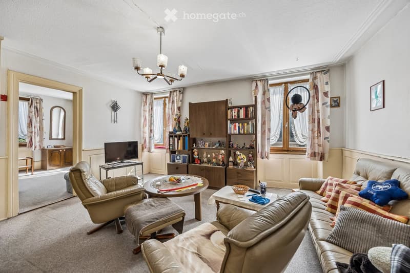 A cozy living room with a beige leather sofa, armchair, coffee table, and bookshelf. The room features large windows with curtains, a chandelier, and a television on a stand.