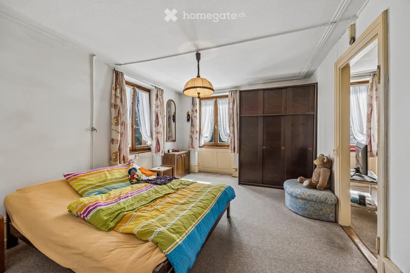 A cozy bedroom with a large bed, colorful bedding, a wooden wardrobe, a small stool with a teddy bear, a hanging lamp, and a mirror on the wall.