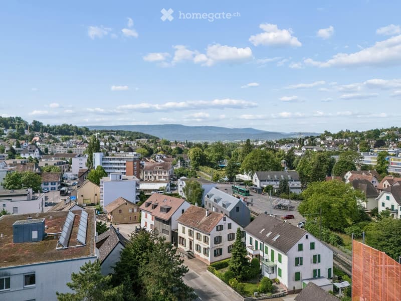 Aerial view of a suburban town with a mix of residential and commercial buildings, surrounded by greenery and distant hills.