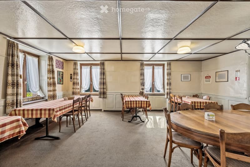 Restaurant or dining area with multiple tables and chairs, patterned tablecloths, large windows with curtains, and framed pictures on the walls.