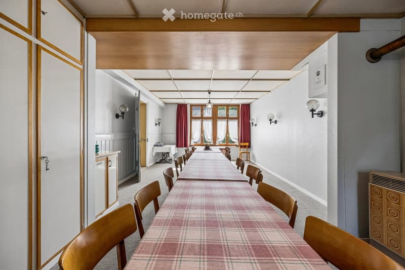 A long, narrow dining area with a long table and multiple chairs, wooden cabinetry, and wall-mounted lamps.