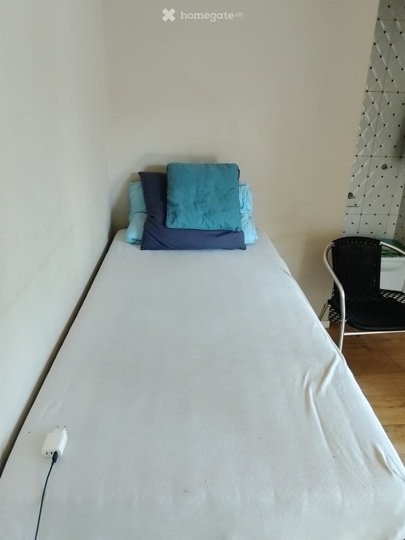 A small bedroom with a single bed, pillows, and a chair in the corner.