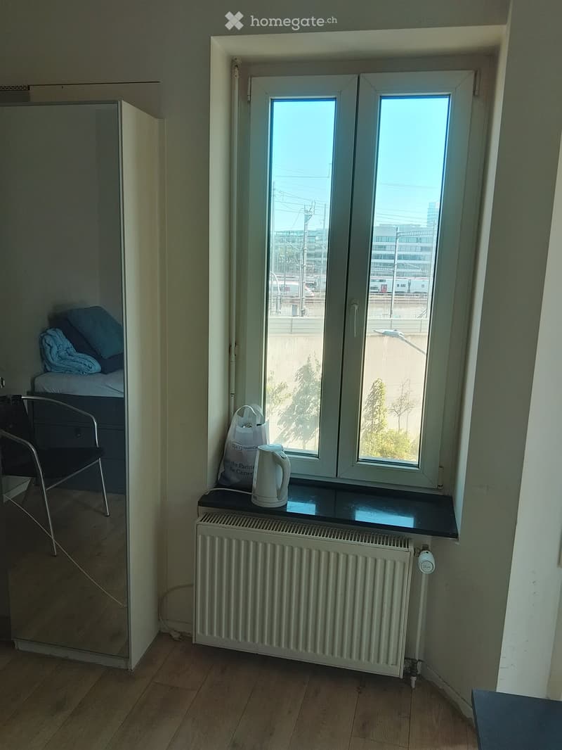 Small furnished room with wooden flooring, a window with a view of the city, a radiator, an electric kettle on the windowsill, a chair, and a bed with pillows.