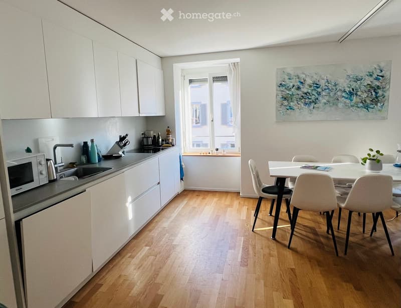 Modern kitchen and dining area with white cabinets, wooden flooring, and a large window. Includes a sink, microwave, oven, dining table, chairs, and a painting on the wall.