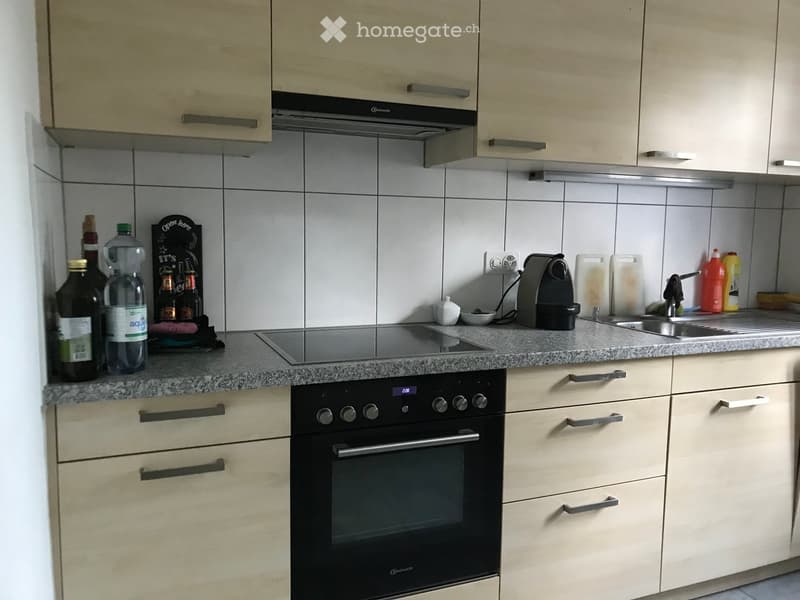 Modern kitchen with light wood cabinets, granite countertop, built-in oven, induction cooktop, sink, and various appliances and items on the counter.