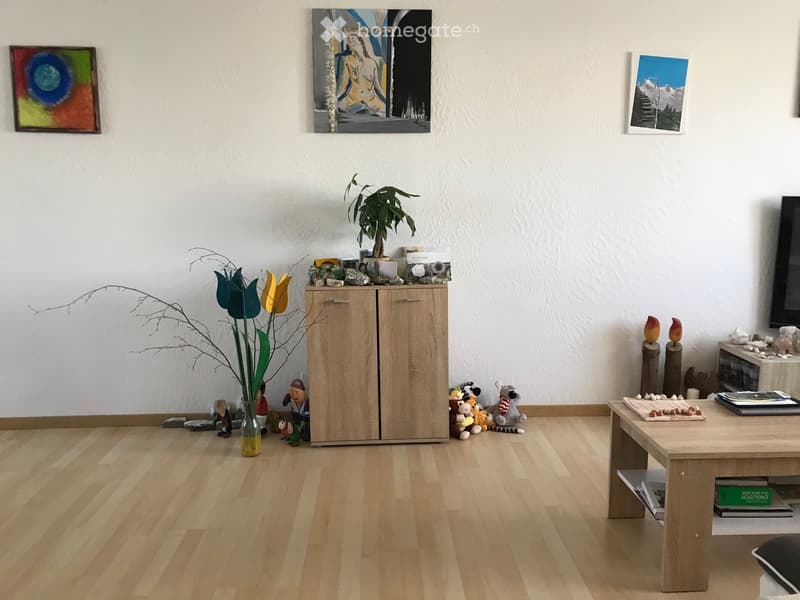 A modern living room with light wooden flooring, a central wooden table, a cabinet, and various decorative items including plants, toys, and artwork on the walls.