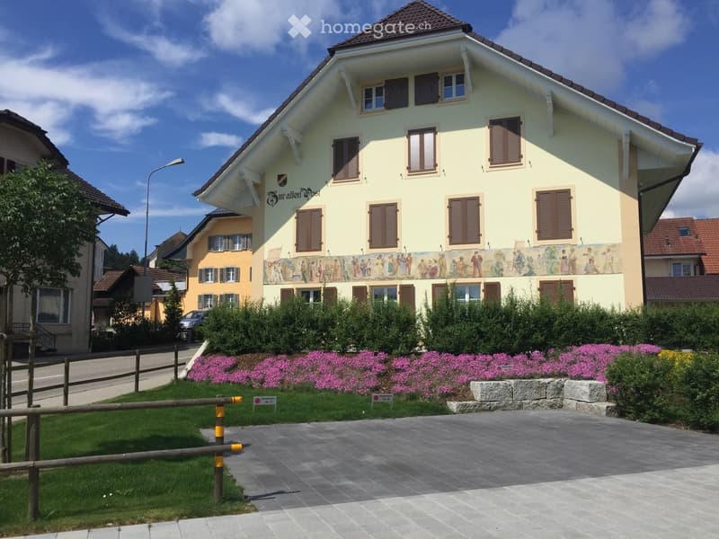 Two-story yellow building with brown shutters, decorative mural, and well-maintained garden with pink flowers and green shrubs.